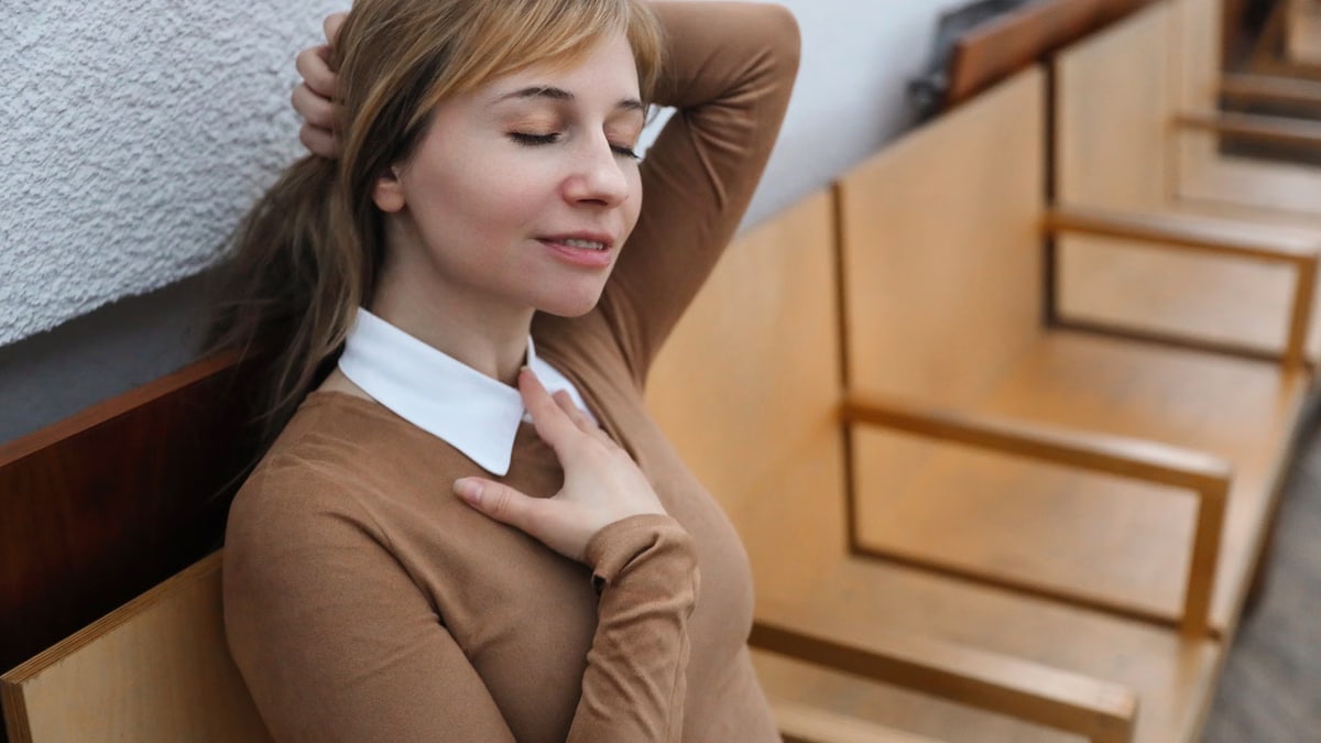 A stylish woman sits comfortably with eyes closed on a wooden bench indoors, capturing serenity.
